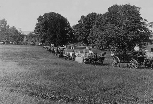 In celebration of Farmers Week 1921, a parade of horse teams and wagons passes a group of observers. Connecticut Agriculture College buildings are visible in the background. (University Photograph Collection, Archives &amp; Special Collections)