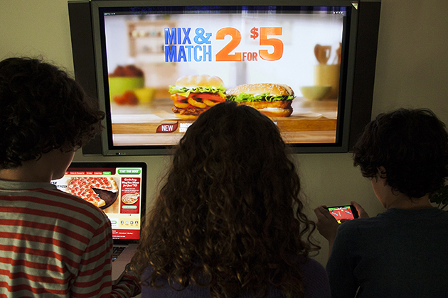 Three minority kids looking at fast food advertising on a television, a laptop, and a smart phone.