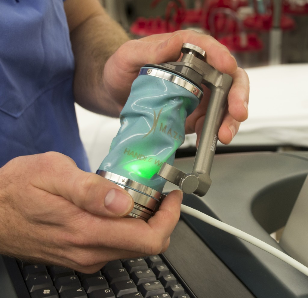 Orthopaedic surgeon Dr. Isaac Moss holds the technology’s small robotic arm, about the size of a soda can, which will help him pinpoint the most precise spot to place screws and other hardware into a patient’s spine. (Janine Gelineau/UConn Health Photo)