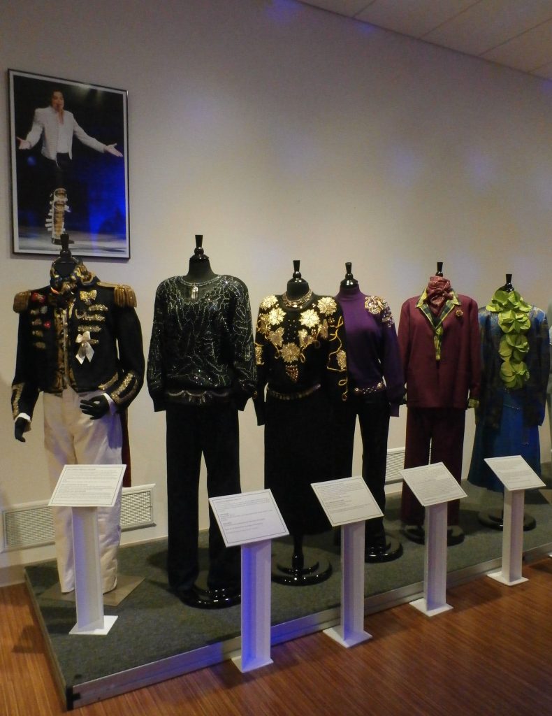 A poster of pop star Michael Jackson attired in a military-influenced stage costume, watches over similarly styled clothing at the Jorgensen Gallery: from left, Civil War cavalry tail coat with embellishments, Versace silk vest and neck scarf, 1986; black ramie and cotton sweater encrusted with iridescent beads and sequins by Priscilla, 1984; sweater with gold applique flowers and leaves by Cedars and knight skirt of lamb’s wool and angora by Outlander, 1983; purple turtle neck sweater, gold embroidery shoulders with large plastic jewels by Ann Tijan, with black, stretch fake leather jeans, 1983; wine striped pantsuit by Christian Dior, 1983; blue patterned silk jacquard jacket, a Versace knockoff, 1986.