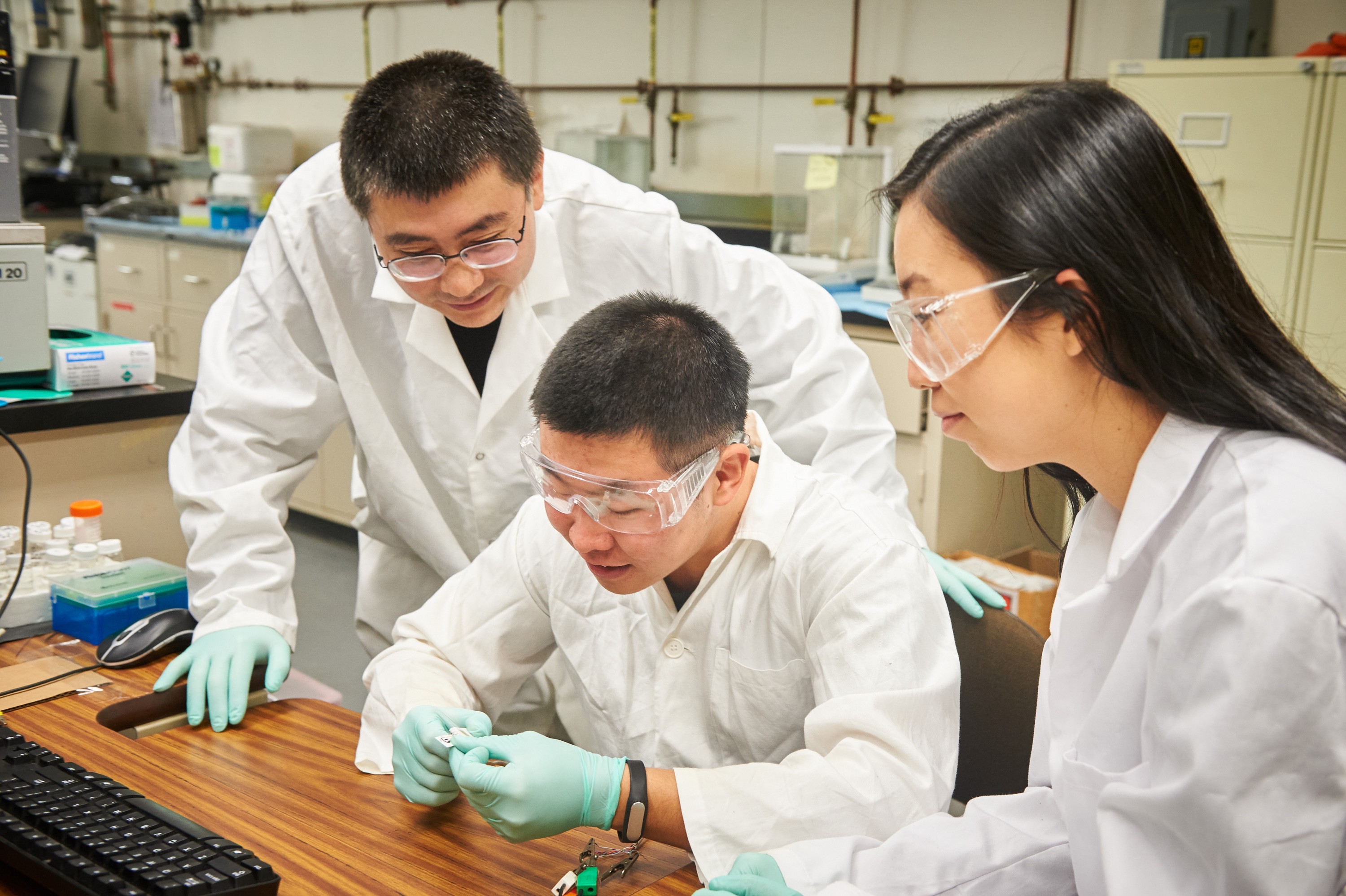 Yu Lei, associate professor of chemical & biomolecular engineering, left, and graduate students Qiuchen Dong and Xiaoyu Ma connect a toxic chemical sensor to a cable in a lab at the United Technologies Engineering Building on Feb. 2, 2016. (Peter Morenus/UConn Photo)