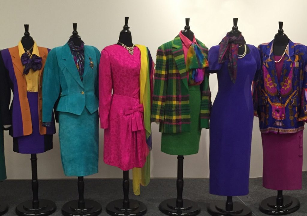 From left, long, color-blocked five-button blazer with gold, purple, and black pieced rayon crepe; matching black and purple skirt by Nichole Miller, 1987; turquoise suede power suit with long ‘80s lapels, with polyester jacquard blouse with jewel-toned paisley overprint by Hastings and Smith, 1985; fuchsia silk jacquard dress by Arnold Scaasi, 1988; green and yellow plaid skirt suit, with pink red and purple mohair, with matching Kelly green tweed skirt by Mary Ann Restivo, 1982; deep violet crepe de chine sheath gown by Maggy of London, 1983;