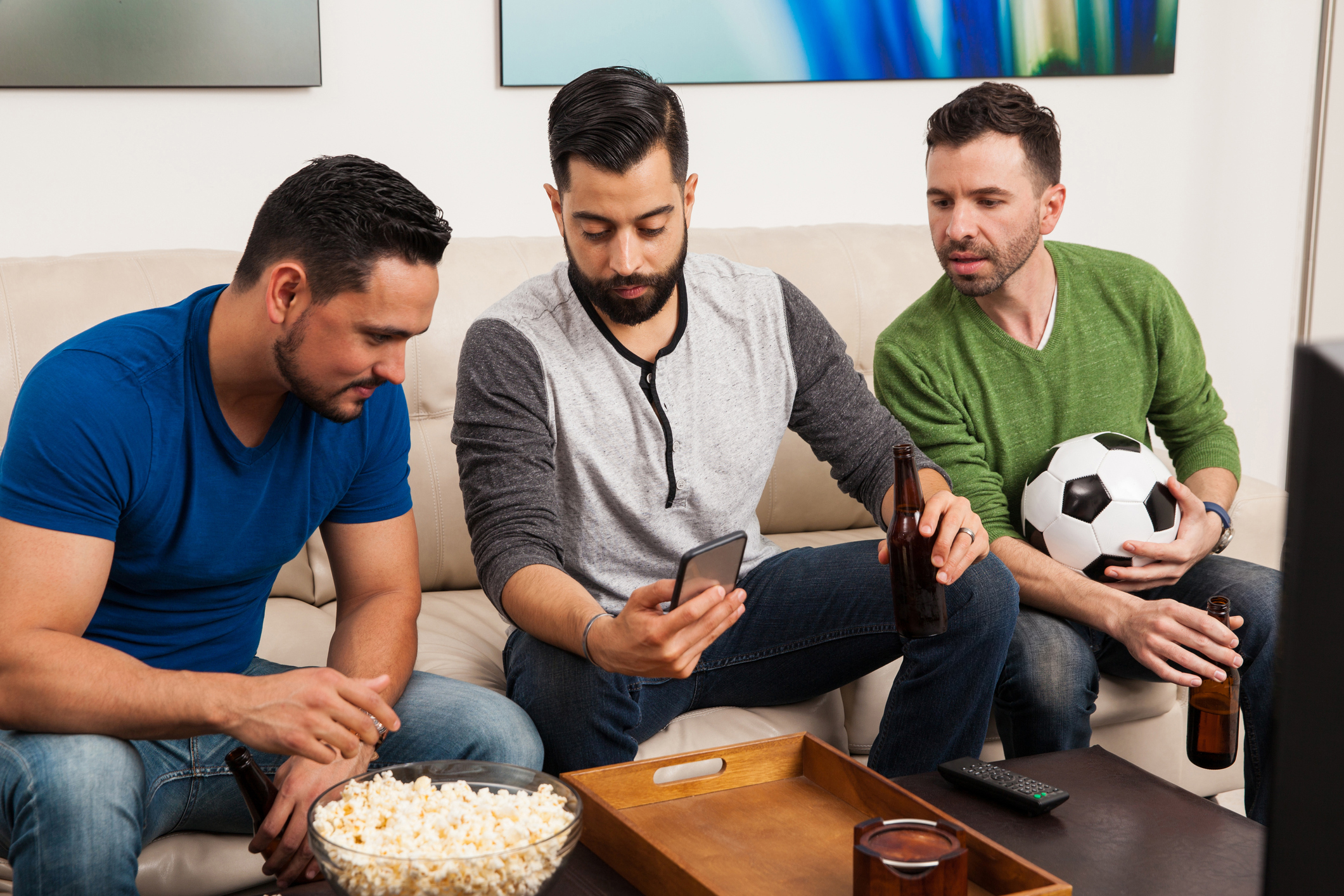 Group of friends checking their team stats on a smartphone while watching a soccer game on TV. (Antonio_Diaz/Getty Images)
