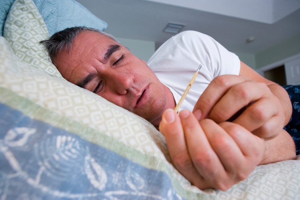 Sick man in bed holding thermometer. (Juan Silva via Getty Images)