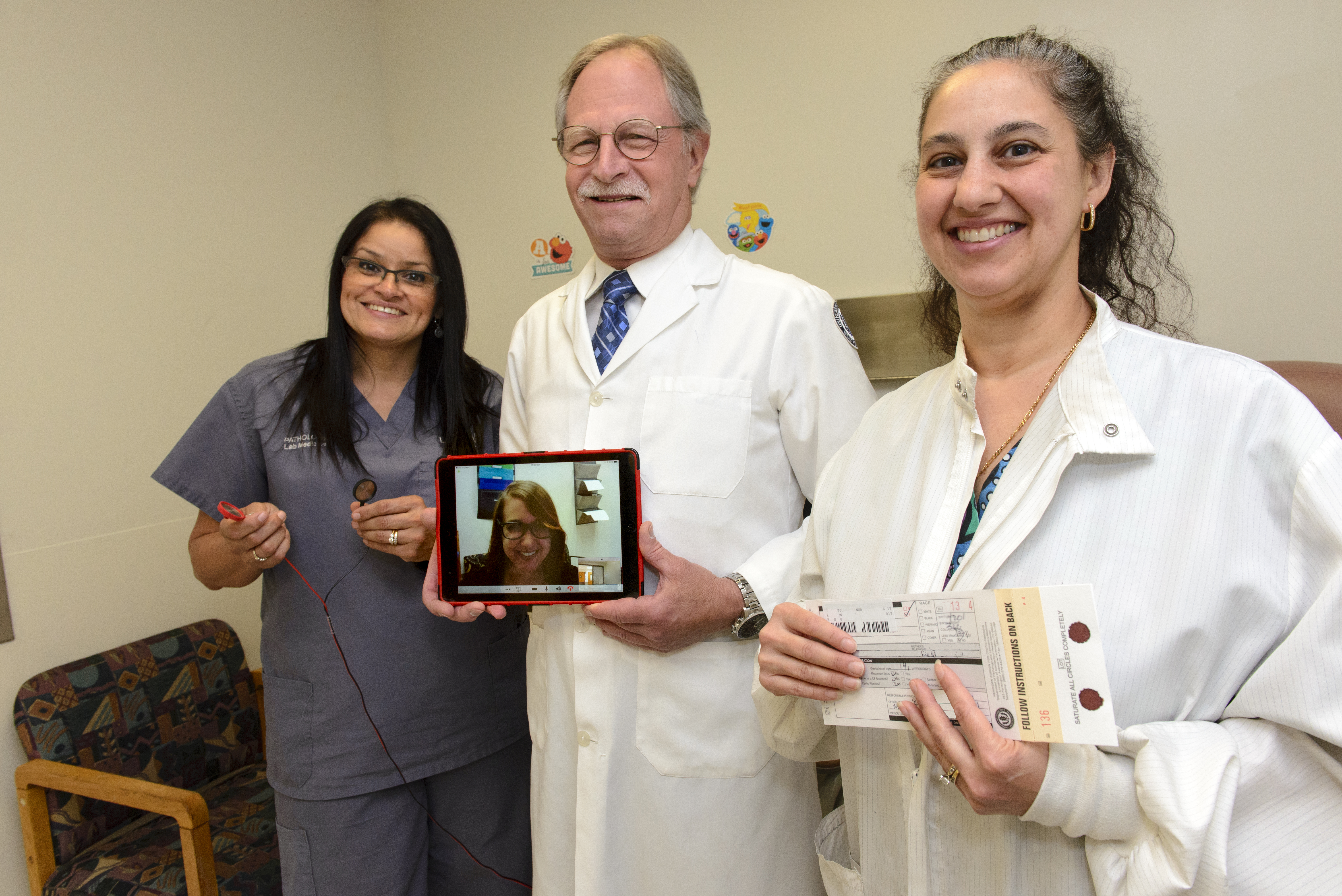 UConn Health's Myra Rivera, Sidney Hopfer, and Giuseppa Santaniello (left to right) demonstrate tools used to screen newborns for cystic fibrosis and provide genetic counseling for parents. On the tablet is Amy Jonasson, a certified genetic counselor from University of Florida Health. (Photo by Janine Gelineau)
