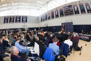 Members of the media and the university community, including several trustees, attended the welcome event. The student-athletes currently on the team are seated against the wall on Hurley's right.