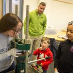 Chemistry major Taryn Wisniewski '19 (CLAS) shows Cyriah Bernard how to press a coin at the chemistry workshop, as Keegan Bowdler and his father Daniel look on in anticipation. (Lucas Voghell '20 (CLAS)/ UConn Photo)