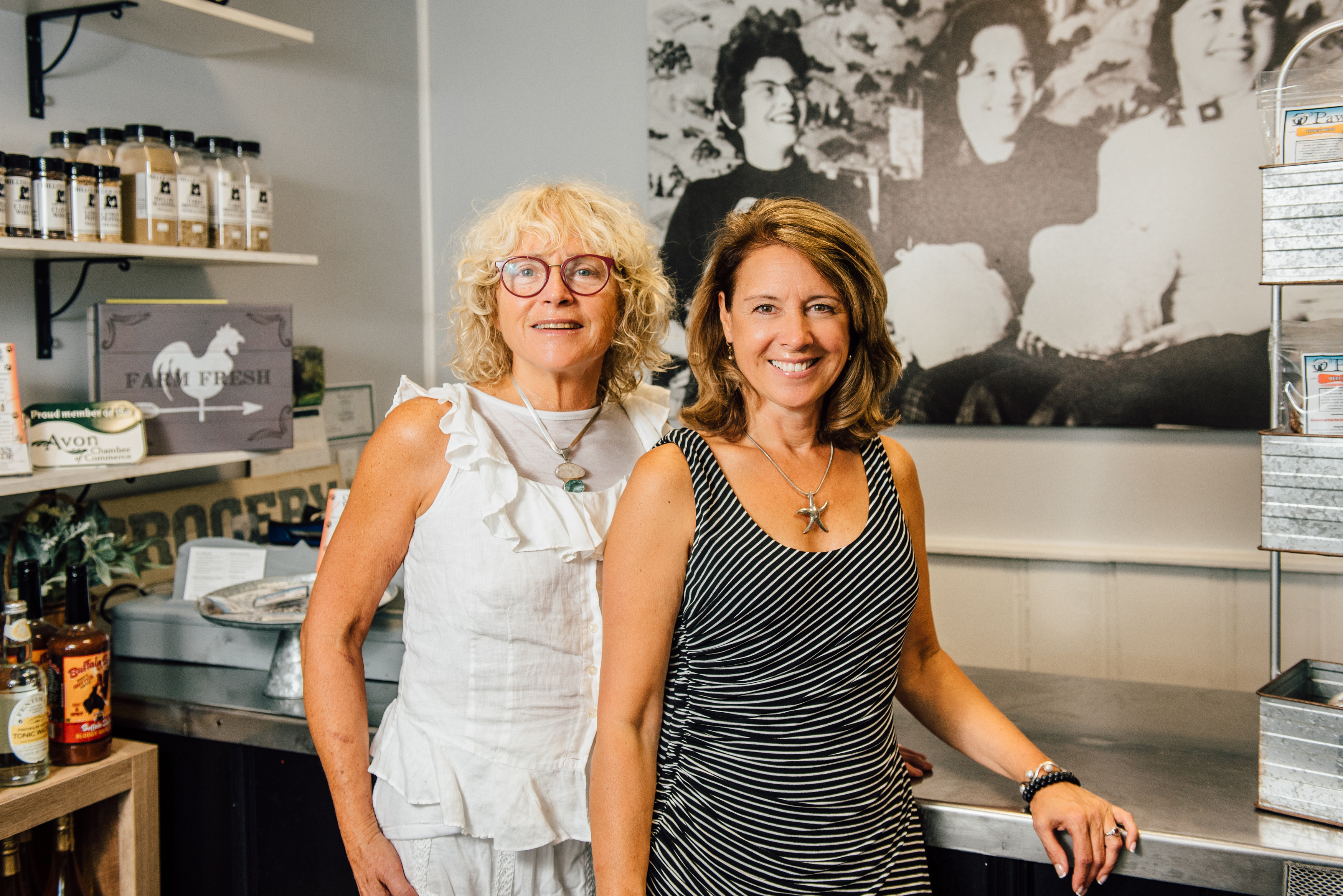 Cal Miller-Stevens, left, and her niece Capri Frank pose for a photo inside the store at Miller Foods Inc., a fourth-generation, family-owned and operated food business located in Avon, Connecticut. Behind them is a photo taken in the early 1960s, in the same location. From left is family matriarch Margaret 'Oma' Miller and her two daughters, Sandi Trudeau (Frank’s mother) and Miller-Stevens. (Nathan Oldham/UConn Photo)