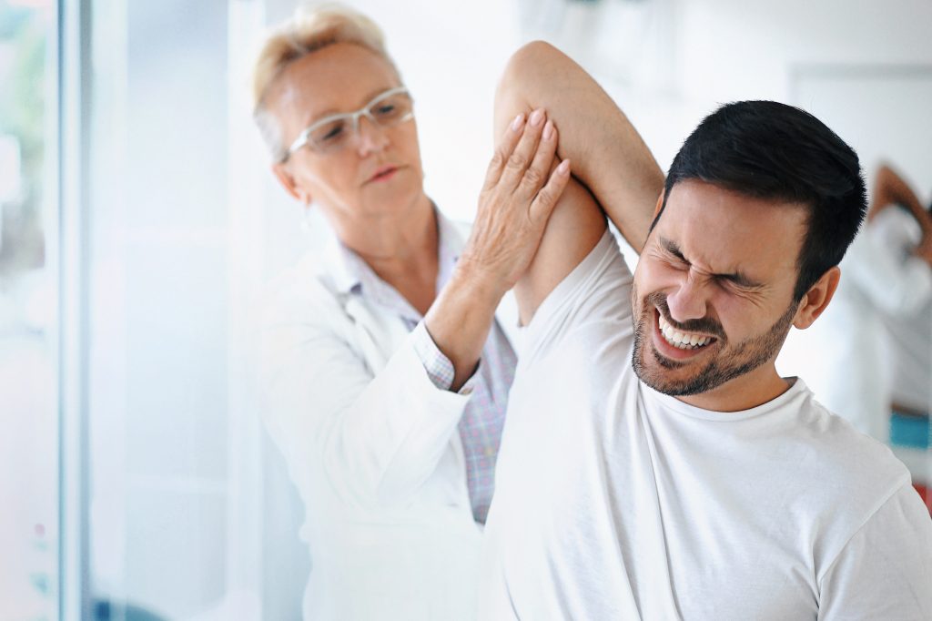 Closeup front angle view of a late 50's female doctor examining a male athlete with some shoulder pain. She's rotating and twisting his shoulder joint and trying to determine which tendons have been damaged. The patient has a painful grimace on his face. (Getty Images)