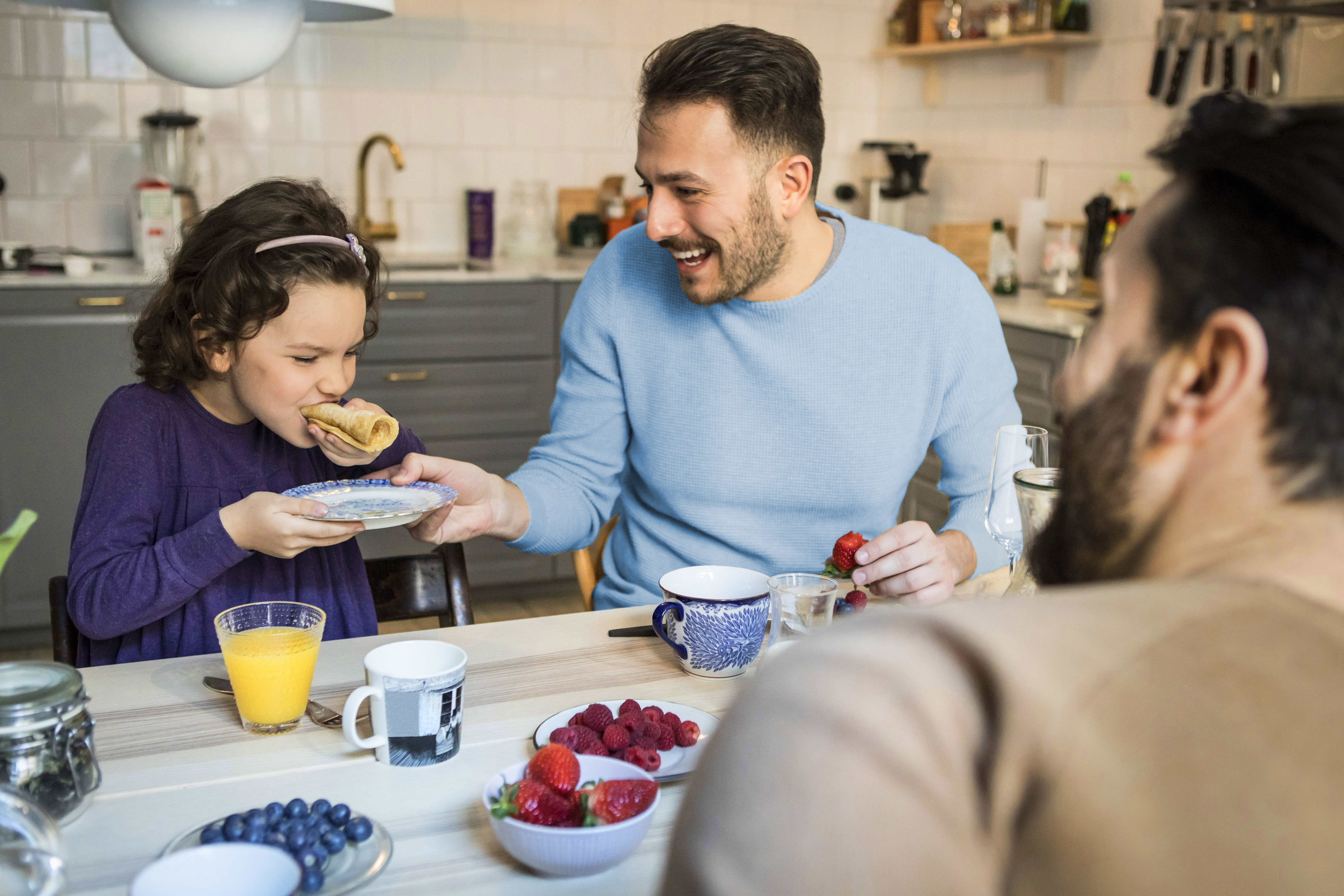 Laughing father holding plate while daughter eats pancake at table. (Maskot/Getty Images)