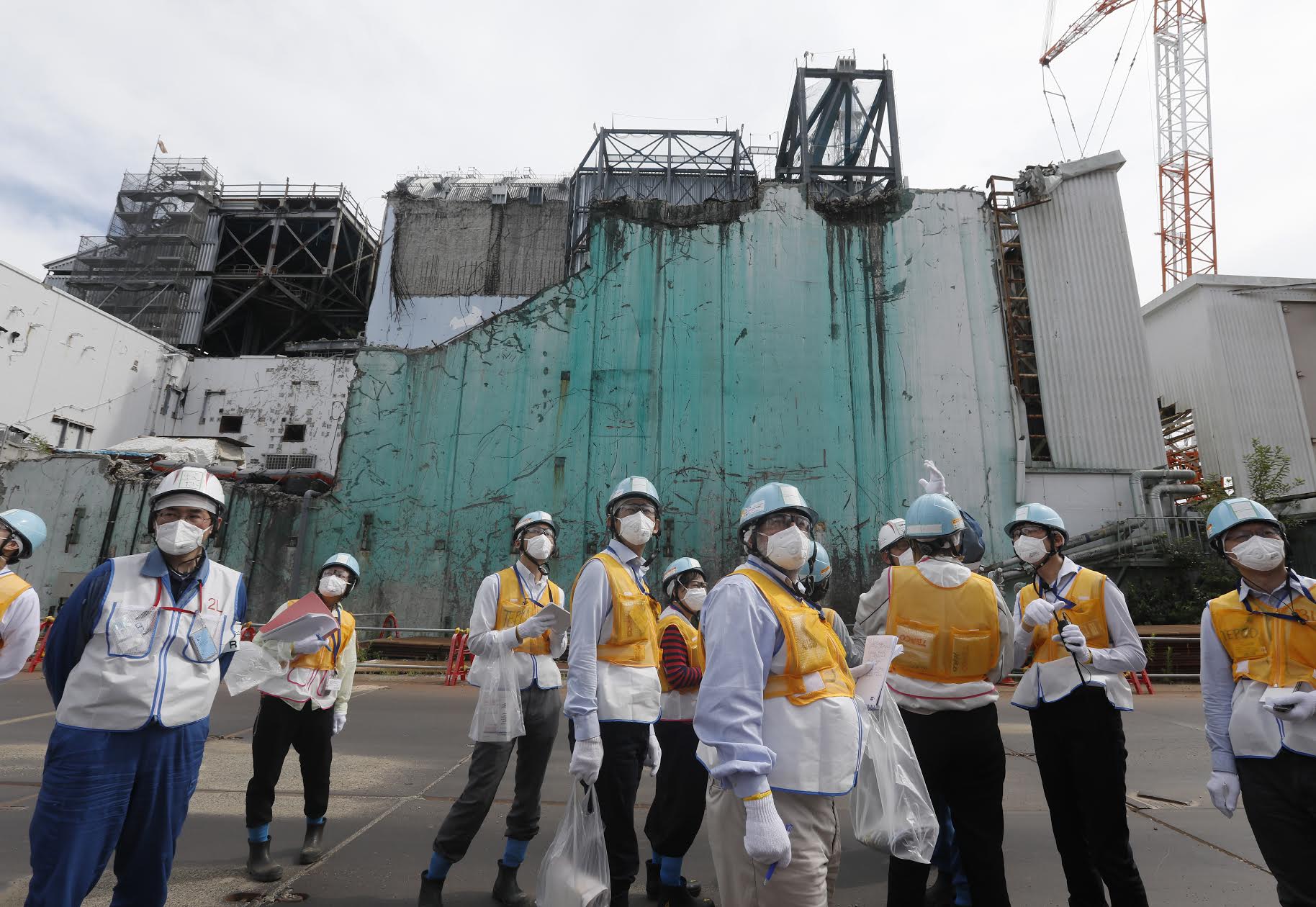 A large group of people wearing safety equipment and breathing masks stands in front of the remains of the destroyed Fukushima nuclear plant in Japan