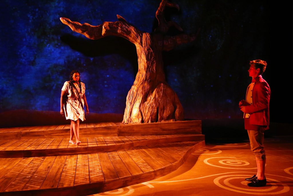 Two actors on stage, an African-American woman and a white man, warily eye each other in a backdrop of Aboriginal Austrlian art and scenery