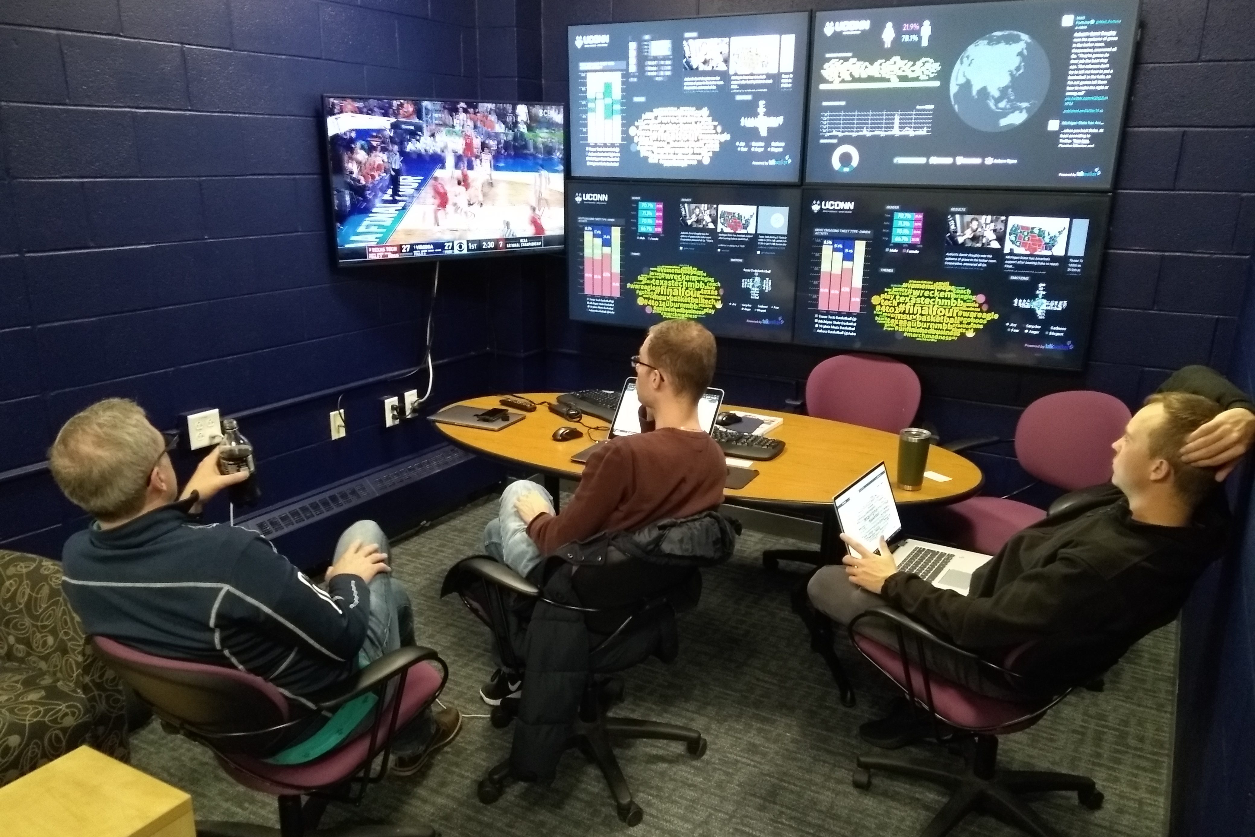 Three men sit in front of a wall full of screens showing a variety of graphics and images relating to social media use during a basketball game.