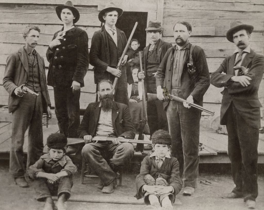 An old photograph shows a group of armed men in front of a rustic cabin