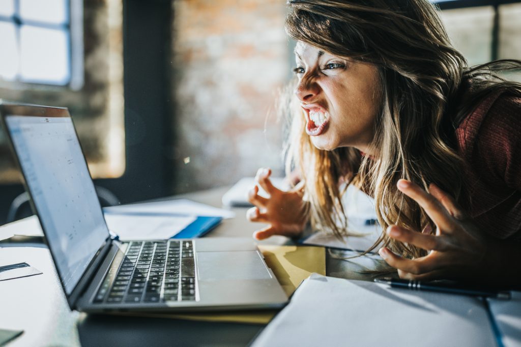 A woman, her face controted in anger, leans over a desk, staring at a laptop computer open in front of her