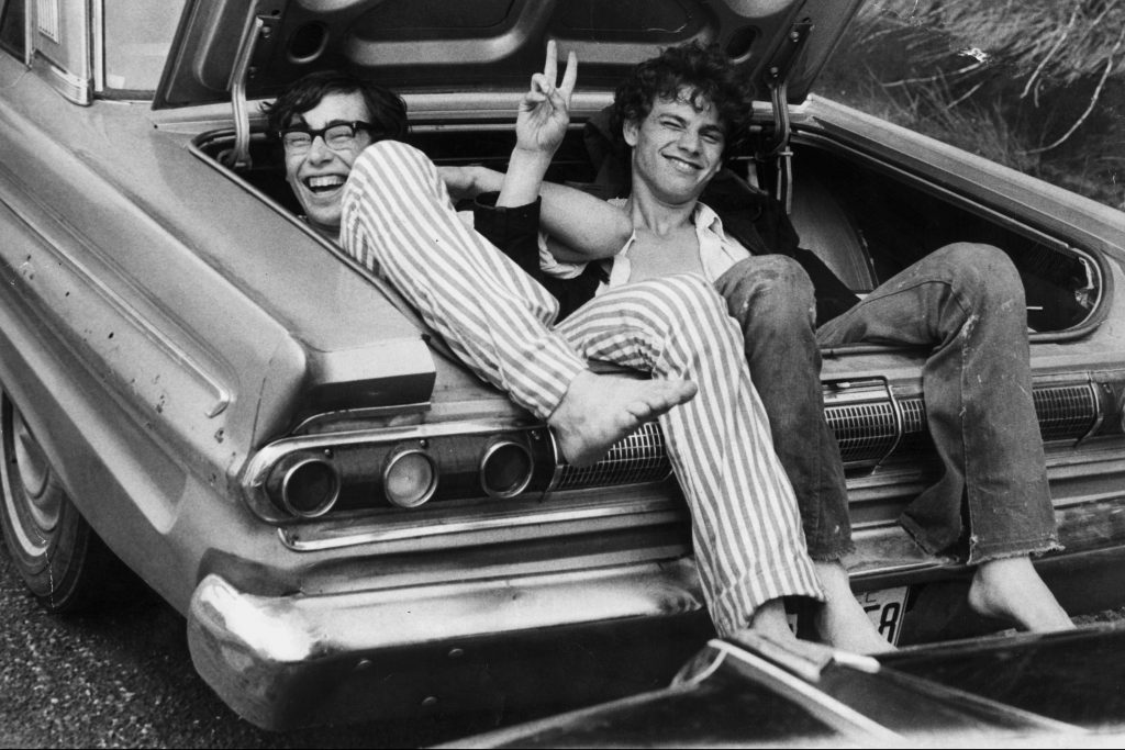 Black and white photo from 1969 of two young men sitting in the trunk of a car, making the peace sign with their hands