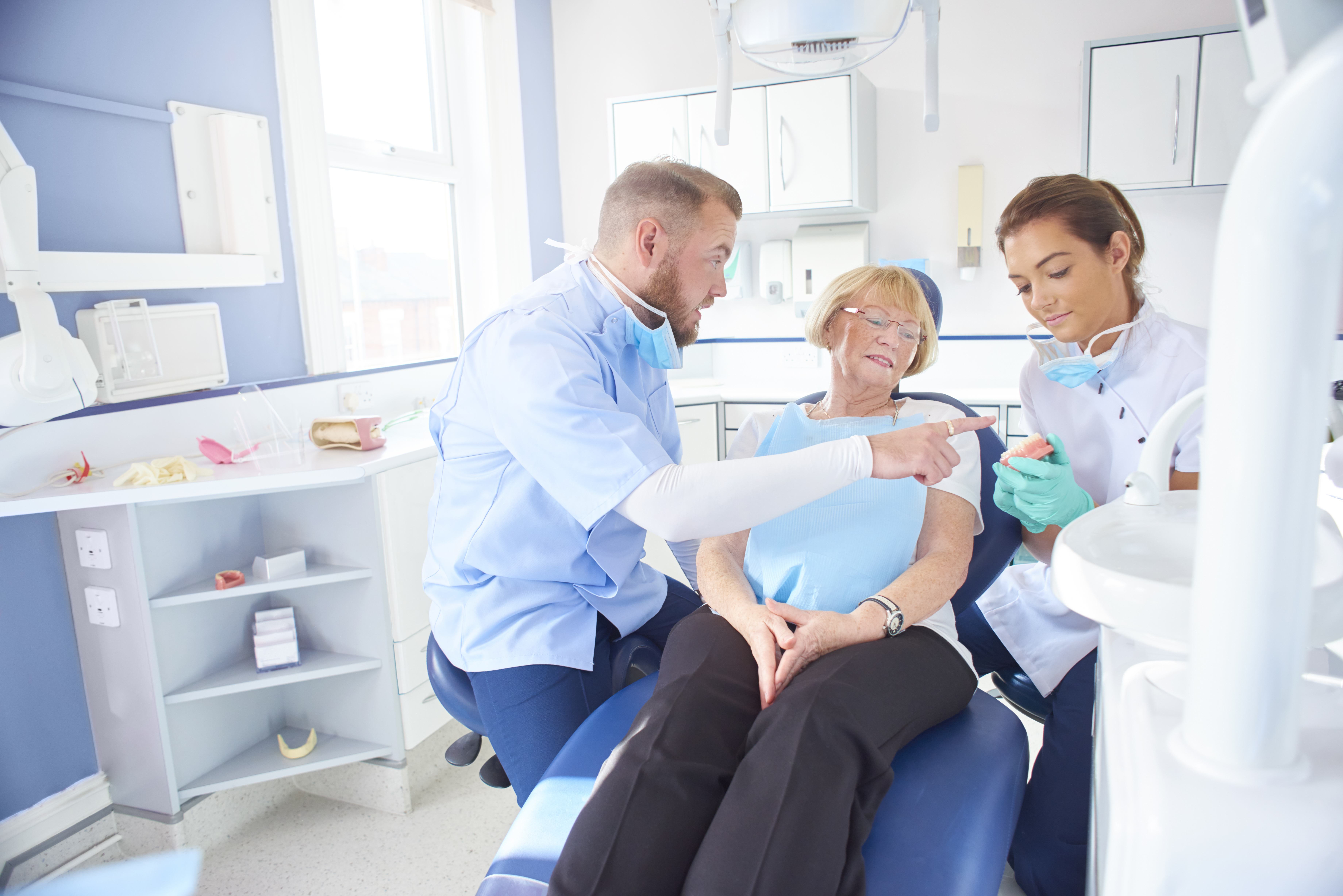 A senior woman is in a dentist's chair , the dentist is showing her where the problem with her teeth is on a set of model dentures which is held by the dental nurse . The brightly lit dentist surgery is clean and modern.