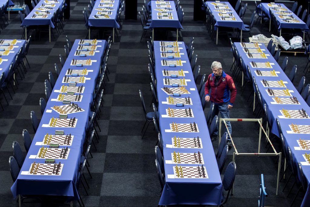 A man with gray hair stands alone in a large room filled with long tables, each one holding many different chess boards