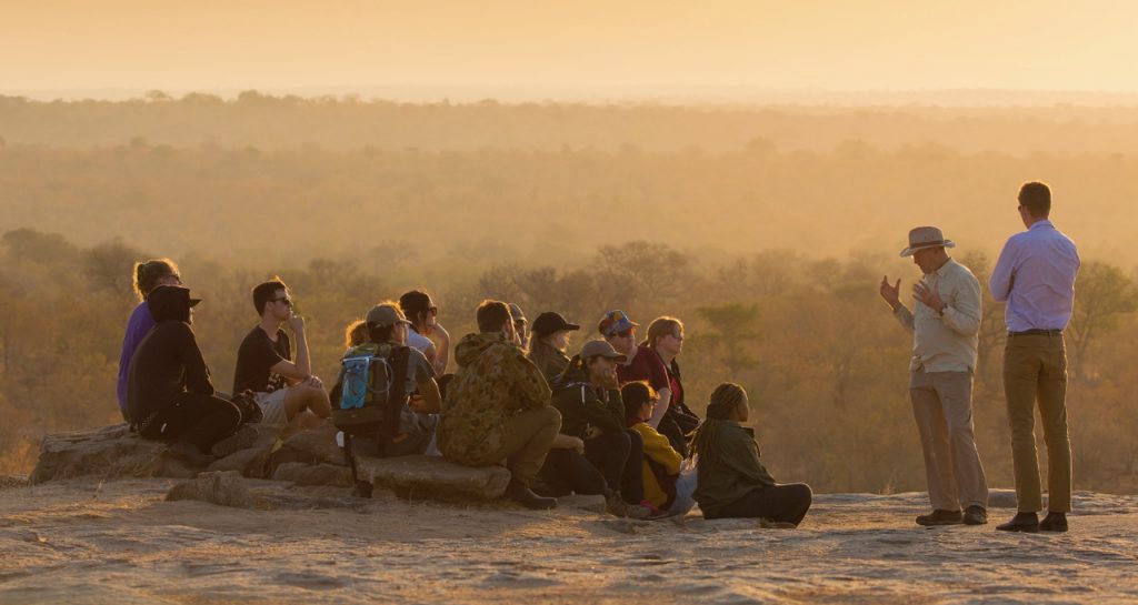 A group of a dozen students sit and two lecturers stand against a South African landscape