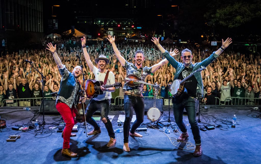 Four men with bluegrass instruments stand on stage before a large audience