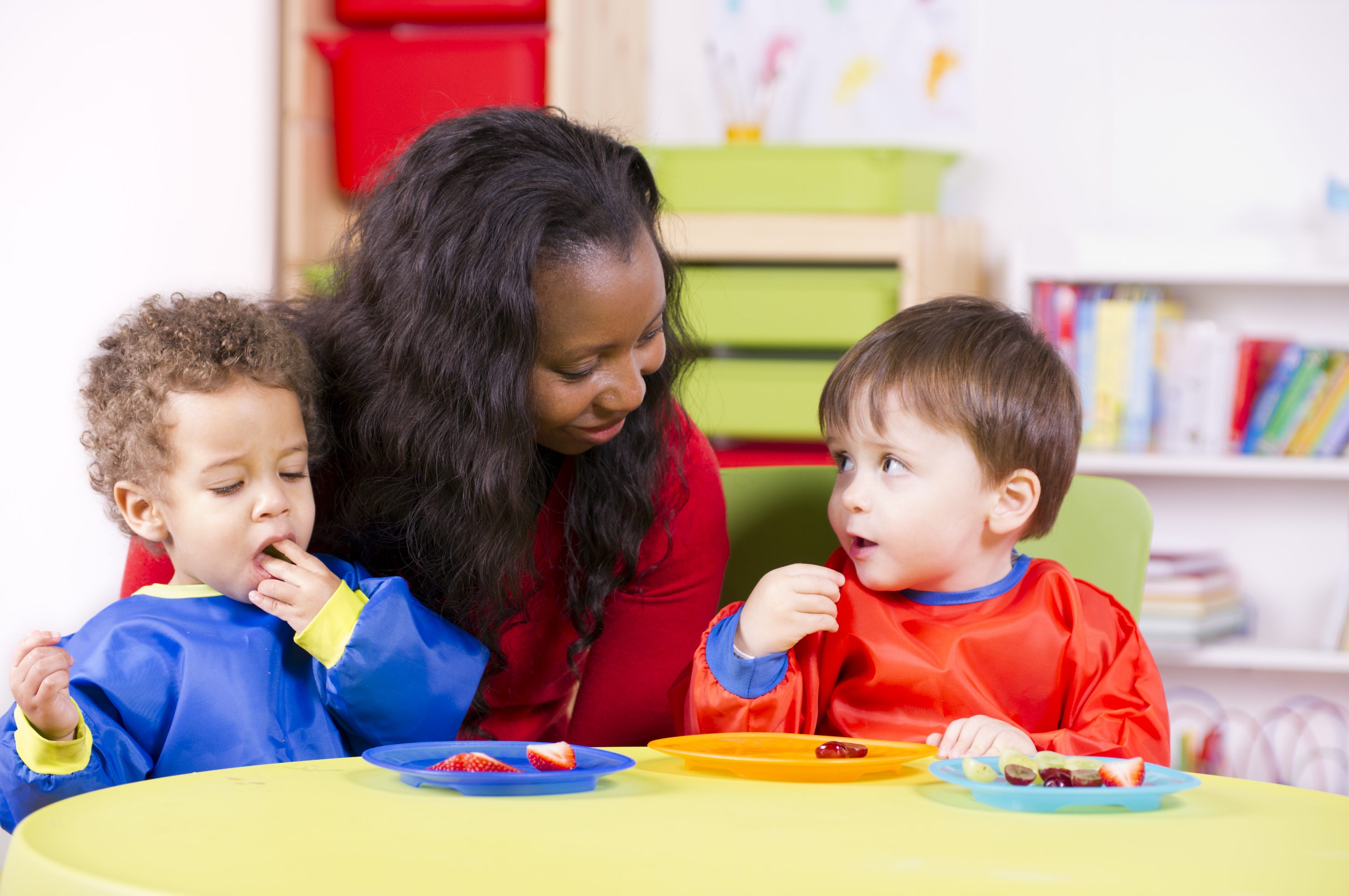 African American educator seated at table with two preschool aged children eating fruit