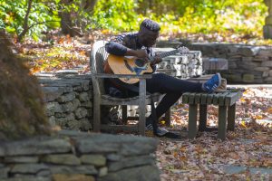 A student playing an acoustic guitar on a stone bench behind the Benton Museum of Art