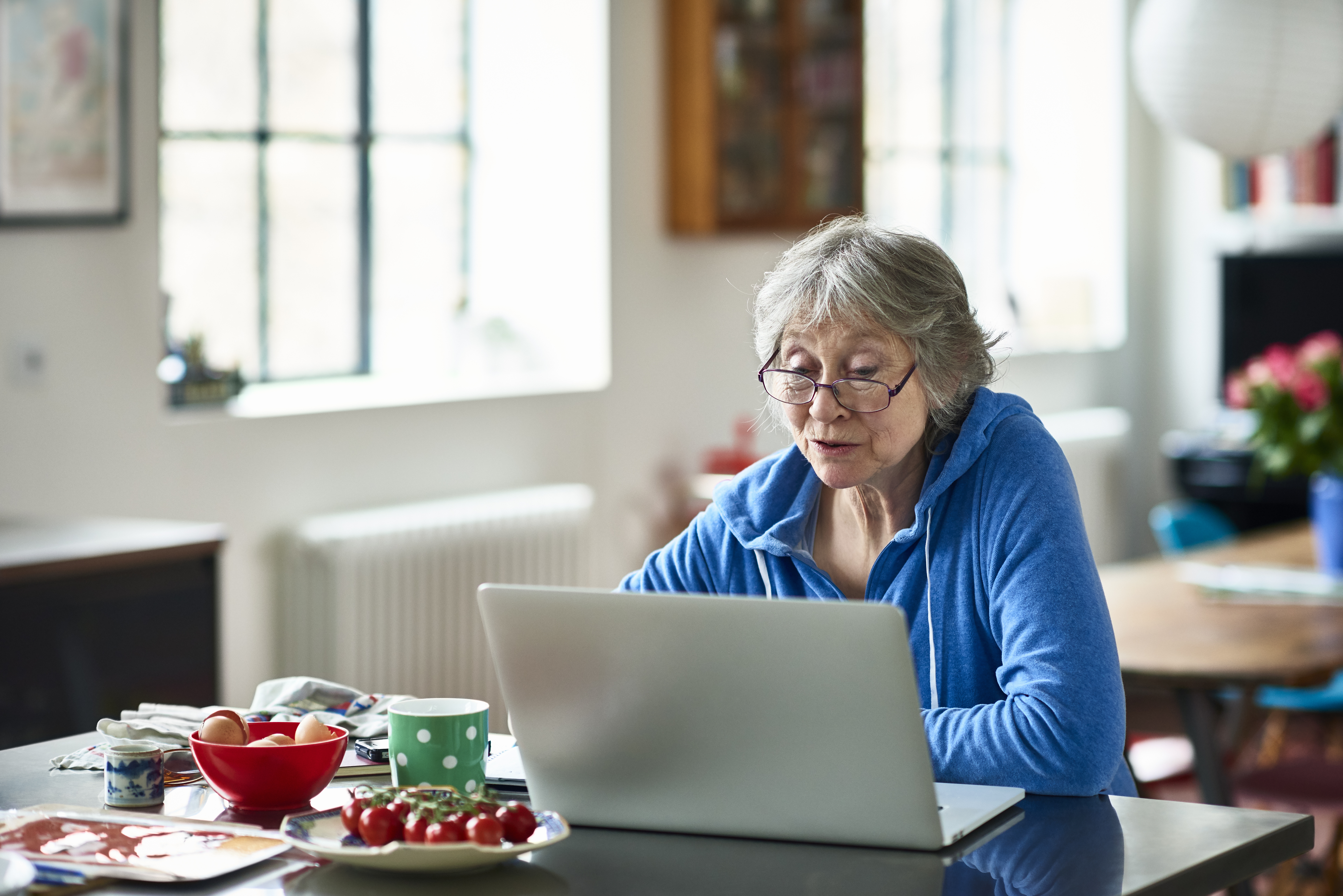 An older woman sitting at a kitchen table, looking at a laptop computer.
