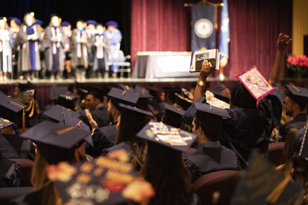 A UConn School of Nursing CEIN/BS graduate, sitting with her peers, facing the stage at the Jorgensen Center, raises her hands to cheer when her campus is announced.