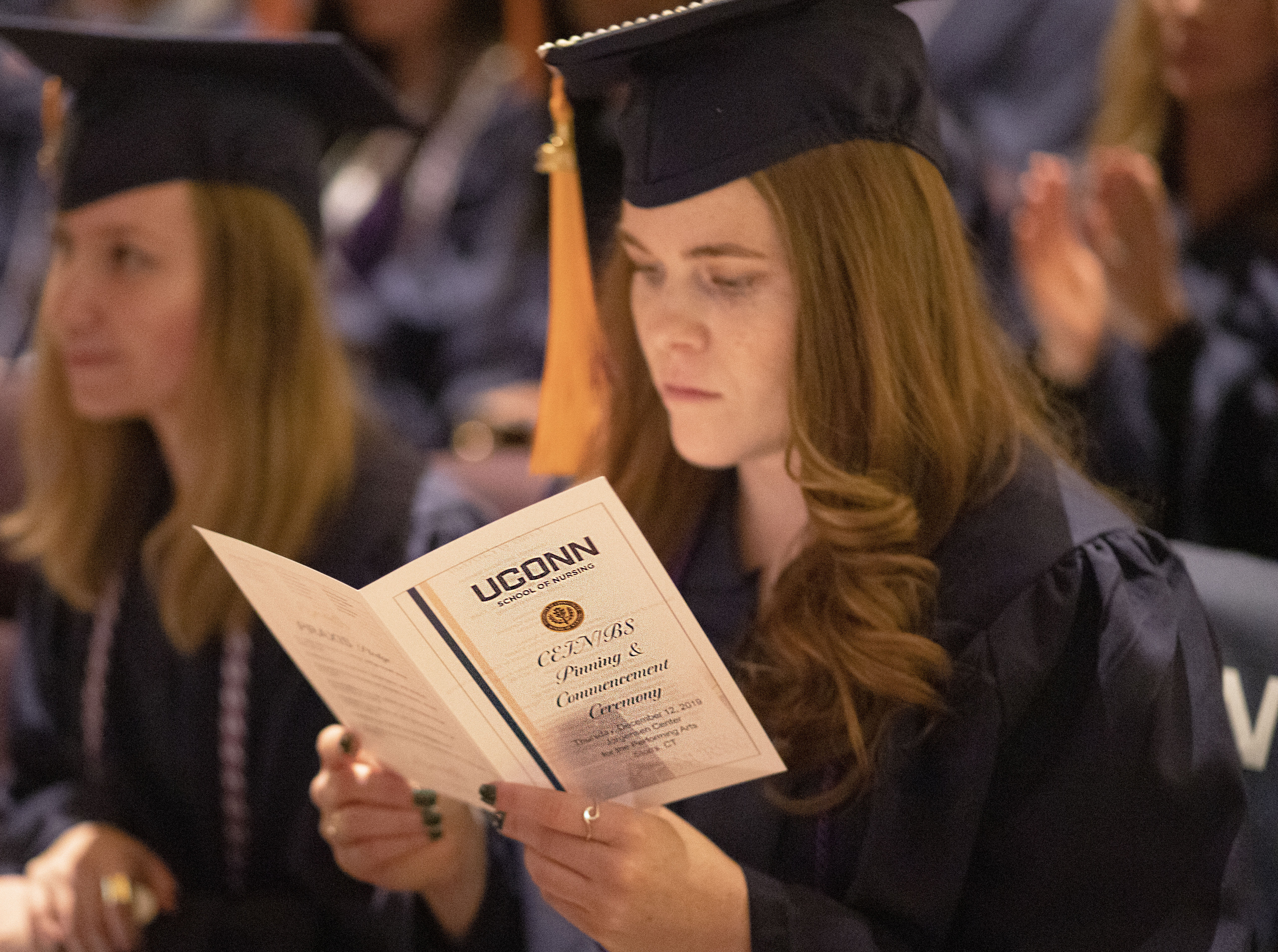 A student, dressed in a commencement cap and gown, reads the program for the UConn School of Nursing's CEIN/BS Pinning and Commencement Ceremony on December 12, 2019