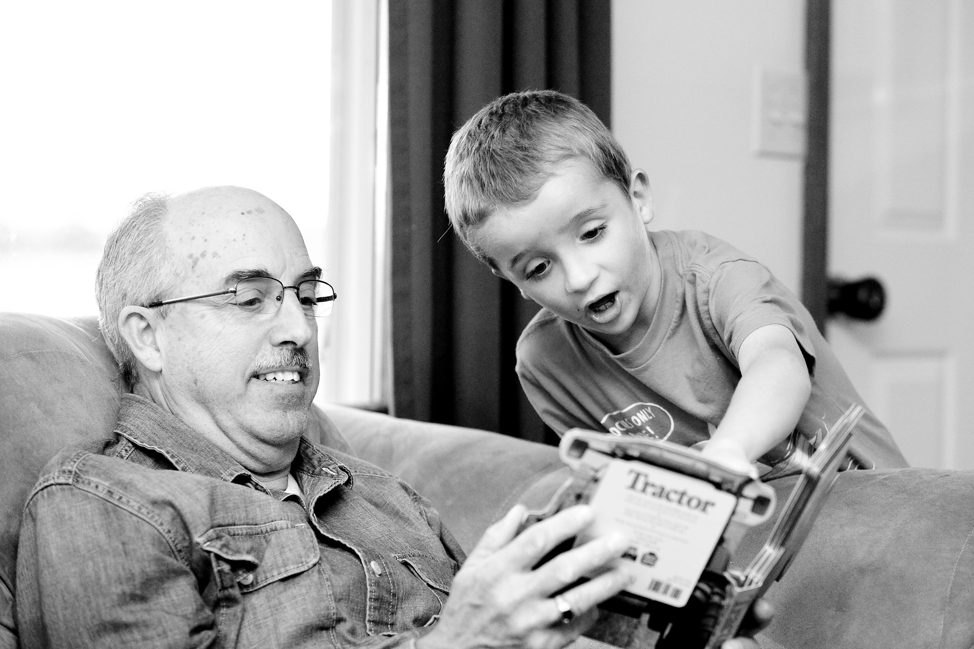 Black and white photo of an older man sitting on a couch holding a book, while a young boy looks on.