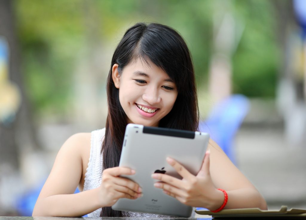 Young asian woman seated outside, smiling, looking at iPad.