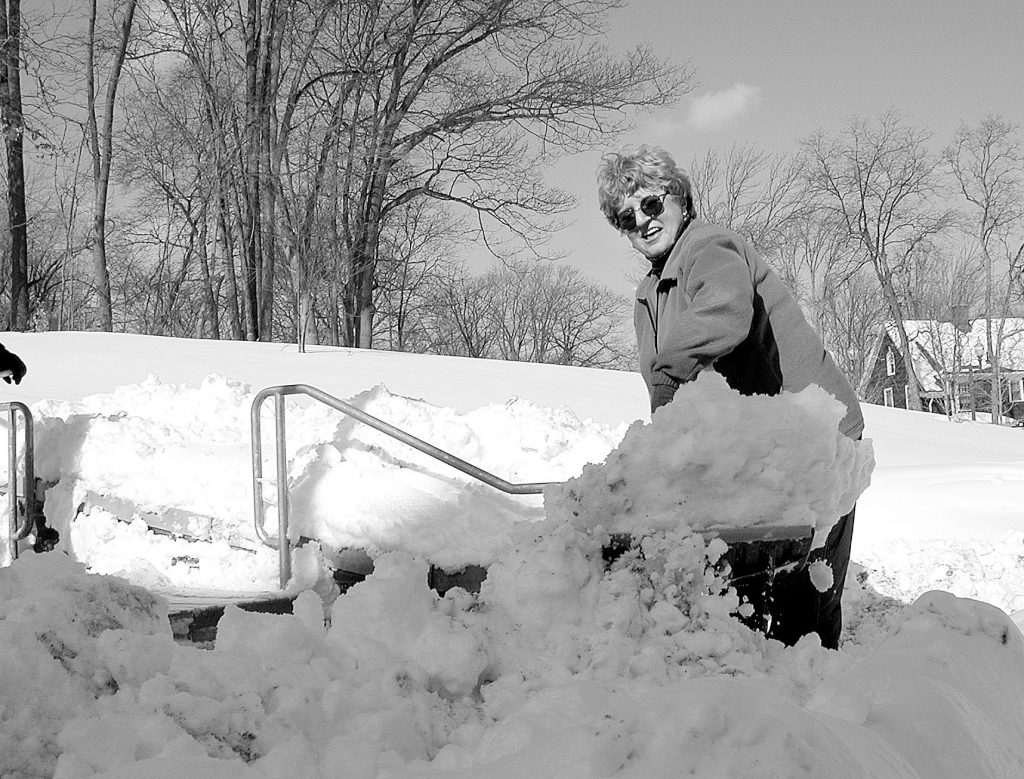 A black and white photo from the 1990s shows a woman shoveling snow on campus.