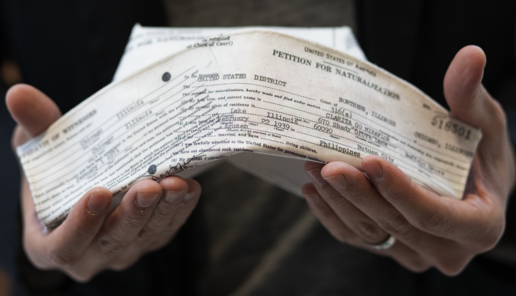 A traditional white nurses' cap is imprinted with the type-written text of an immigration and naturalization form. It it held in a man's hands in this close-up image.