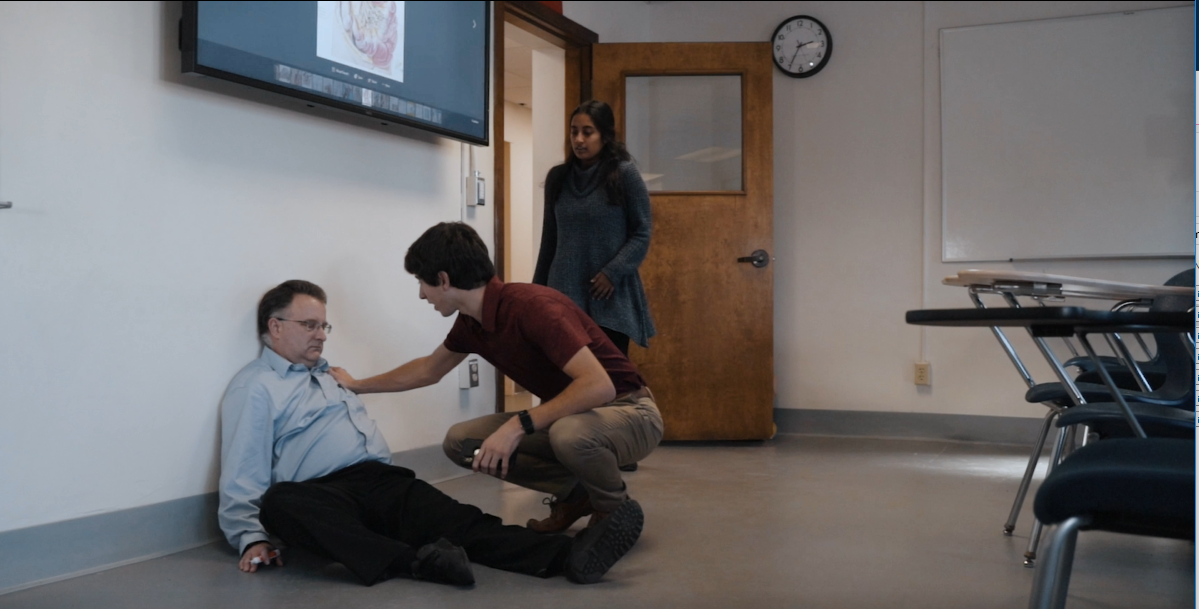 In a still from a training video, students discover an unconscious man sitting on the floor.