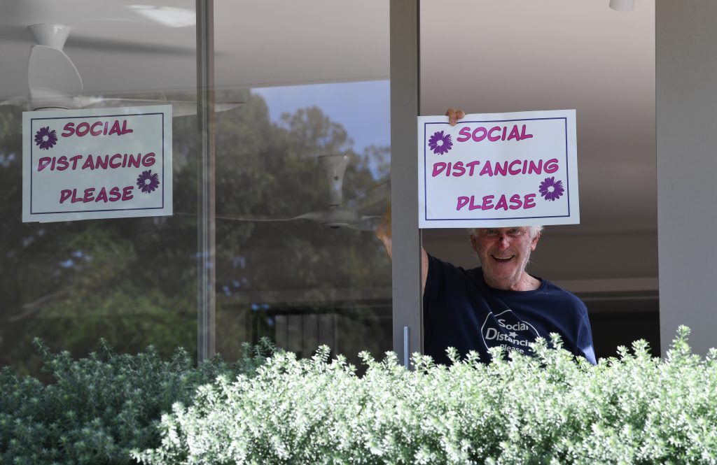 A man inside a house holds up a hand-drawn sign that says "Social Distancing Please."
