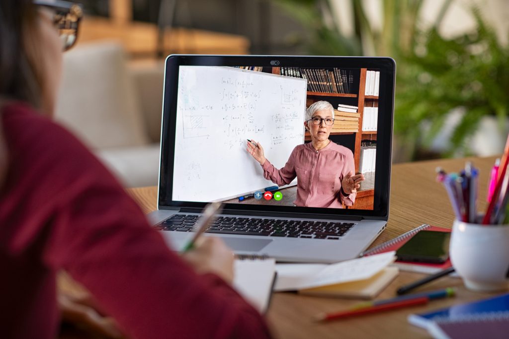 Young student watching lesson online and studying from home. Young woman taking notes while looking at computer screen following professor doing math on video call. Latin girl student studying from home and watching teacher explaining math formula on video chat.