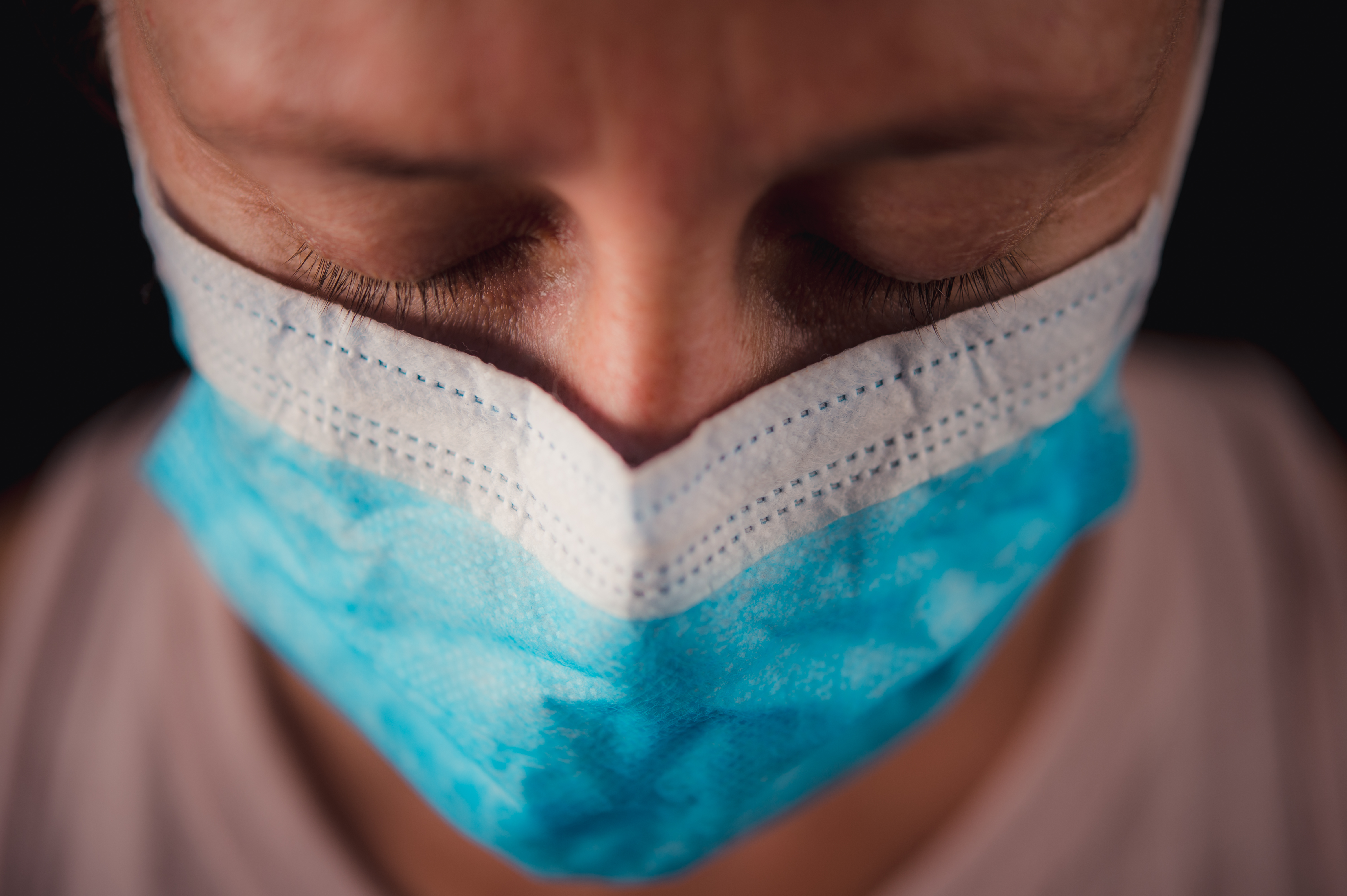 Surgical Mask Close-up headshot portrait with a black background. A beautiful 40-44-year-old woman looking down close-up of her eyelids. Her right eye is wet from running tears. She is sad and emotionally distressed.
