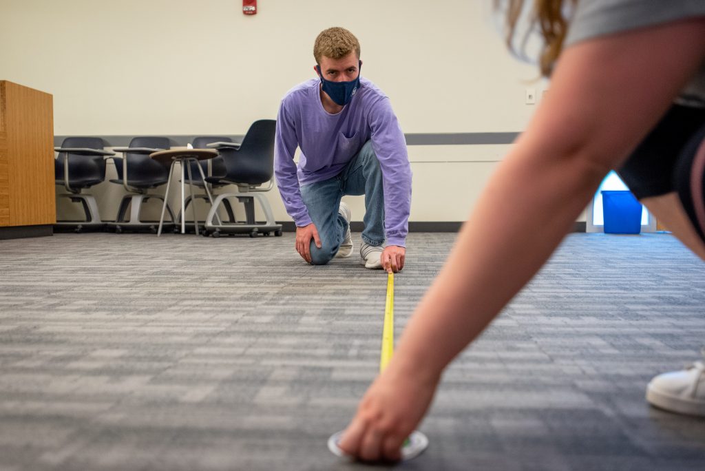 Two people in masks use a tape measure in a classroom to mark off space needed between desks.