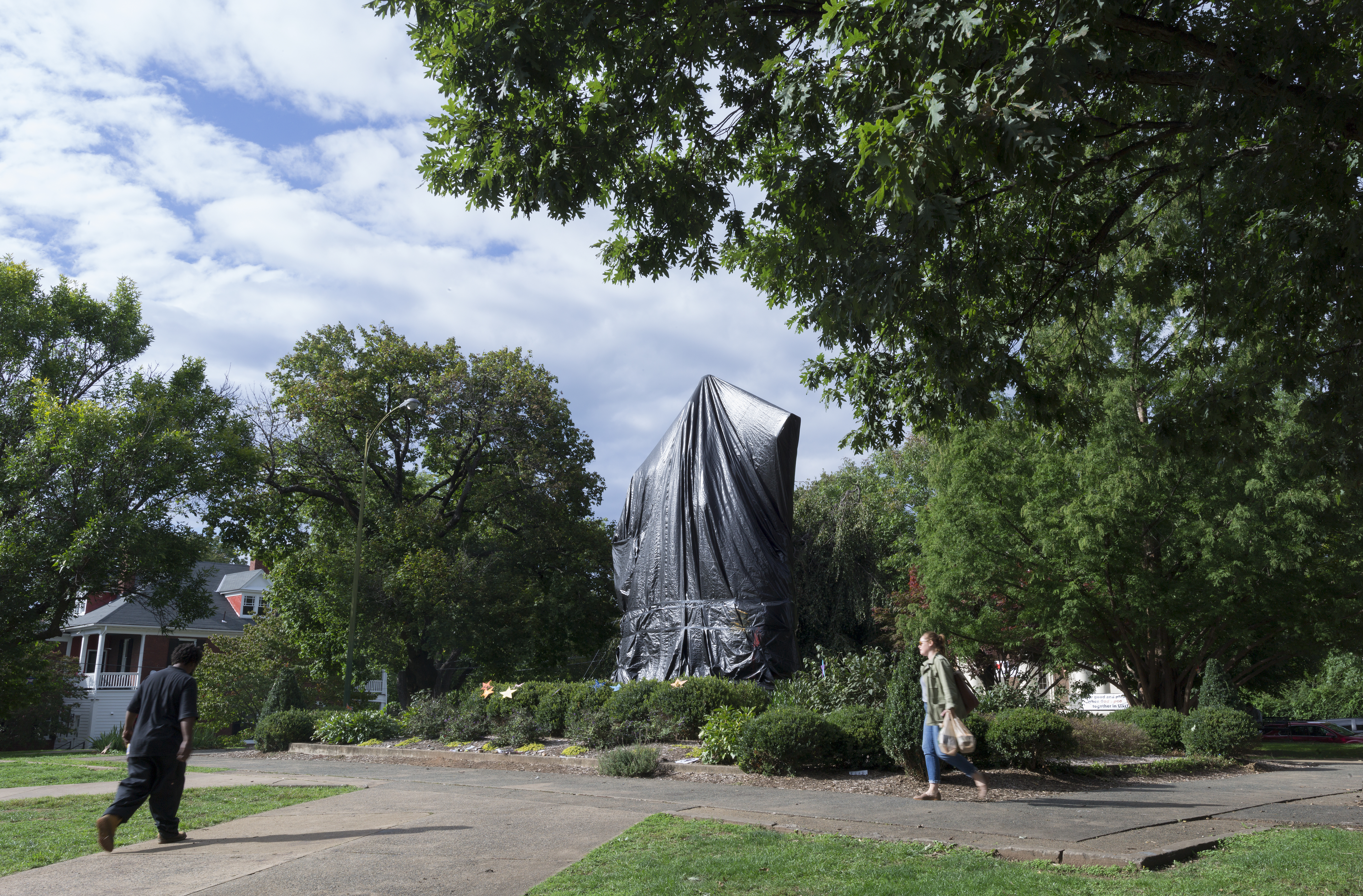Two people in a park walk past a statue of Robert E. Lee entirely covered by a black tarp.