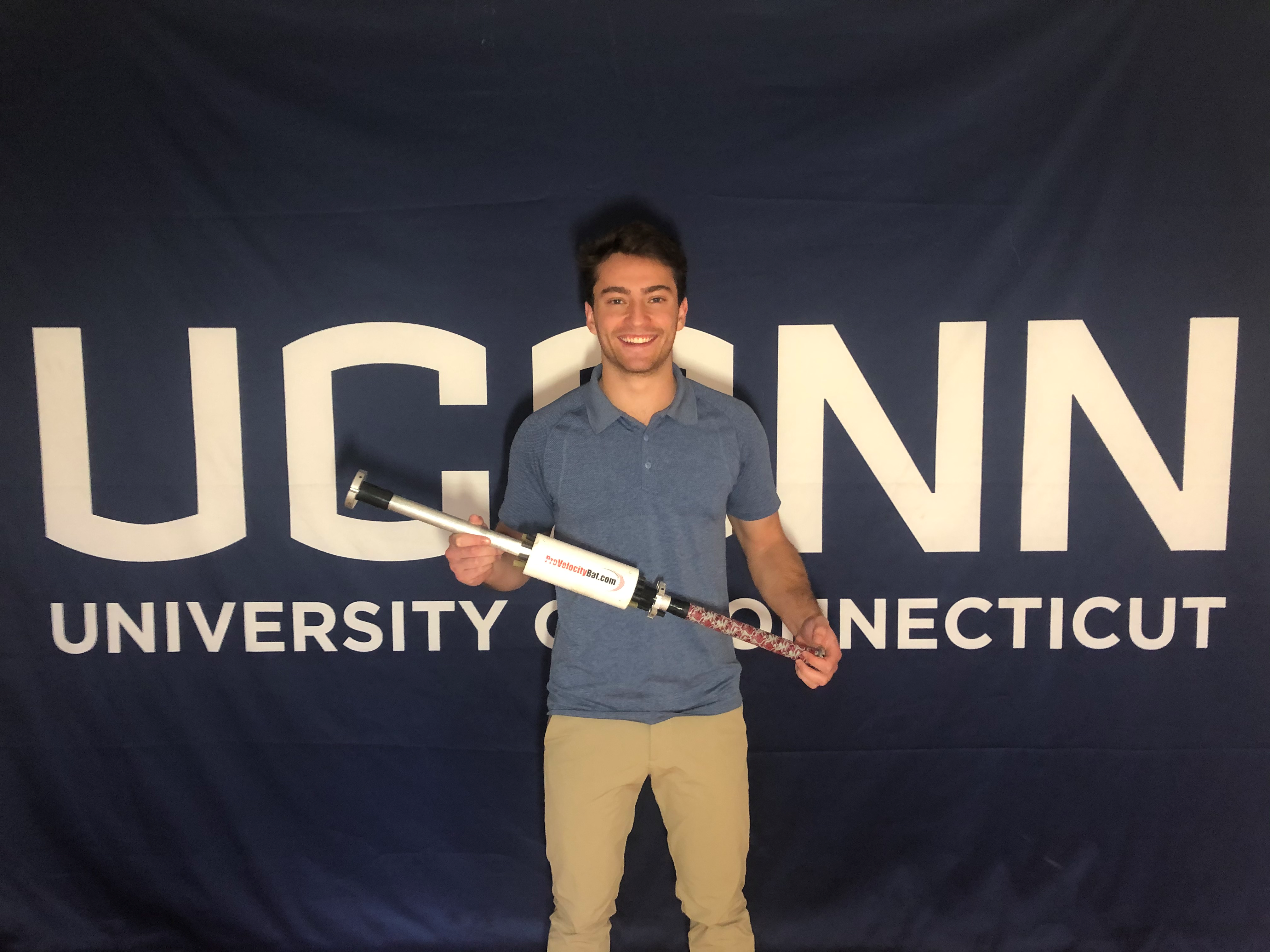 Student entrepreneur Elijah Taitel holds his invention, the Velocity Pro Bat, in front of a UConn banner.
