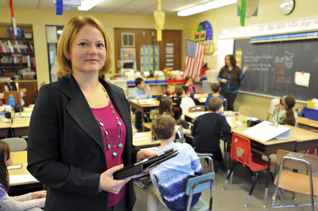 woman in a blazer holding an ipad looks at the camera with a classroom behind her