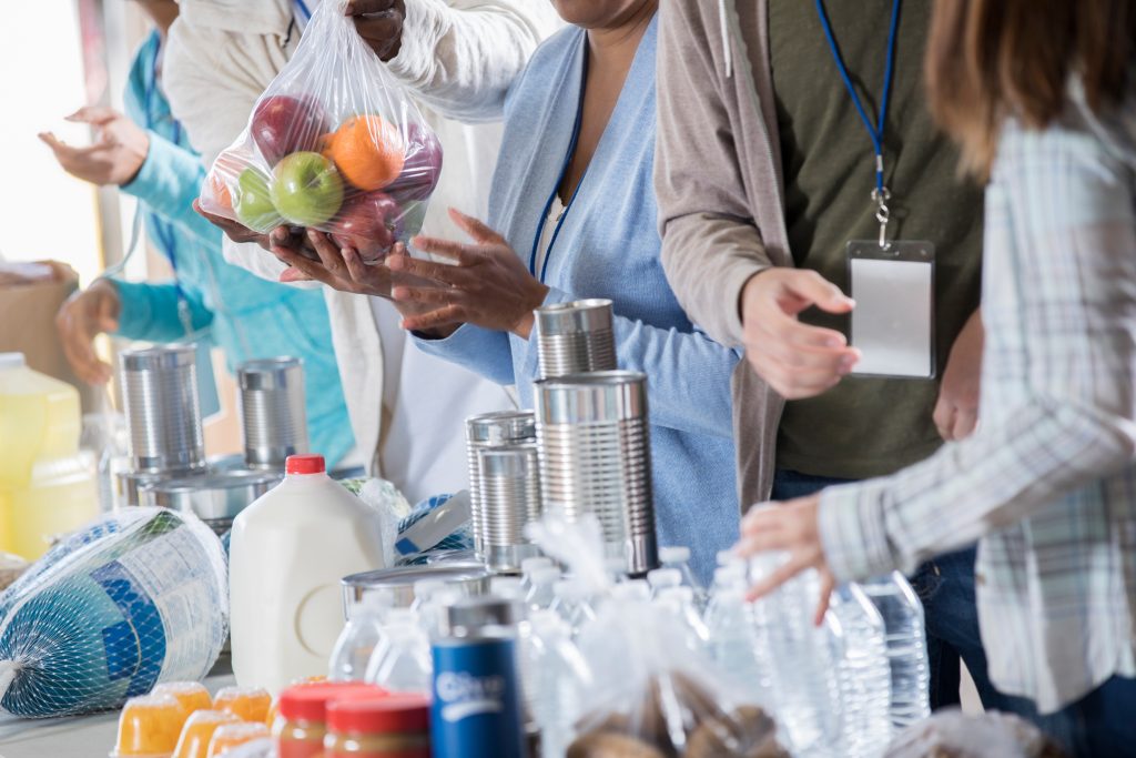 Group of unrecognizable people volunteer at a food bank. They are receiving donated canned food, fruit, milk and other food staples.