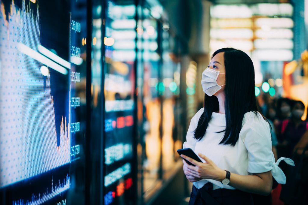 Economic and financial impact during the Covid-19 health crisis deepens. Businesswoman with protective face mask checking financial trading data on smartphone by the stock exchange market display screen board in downtown financial district showing stock market crash sell-off in red colour