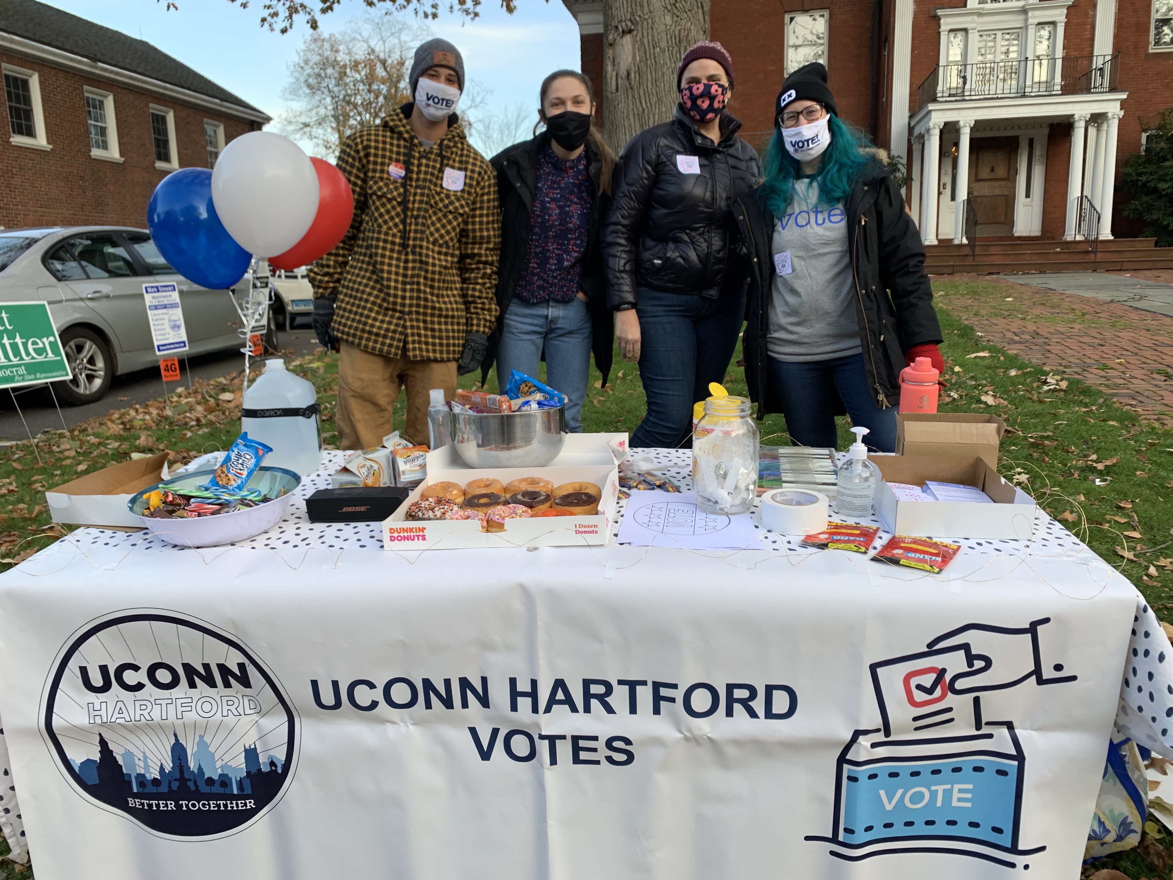 Four people wearing masks stand behind a table with a "UConn Hartford Votes" tablecloth