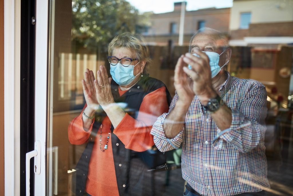 Senior couple in self isolation looking through the window at home clapping. They are showing their support for all the workers and helpers who are helping during the COVID-19 outbreak.