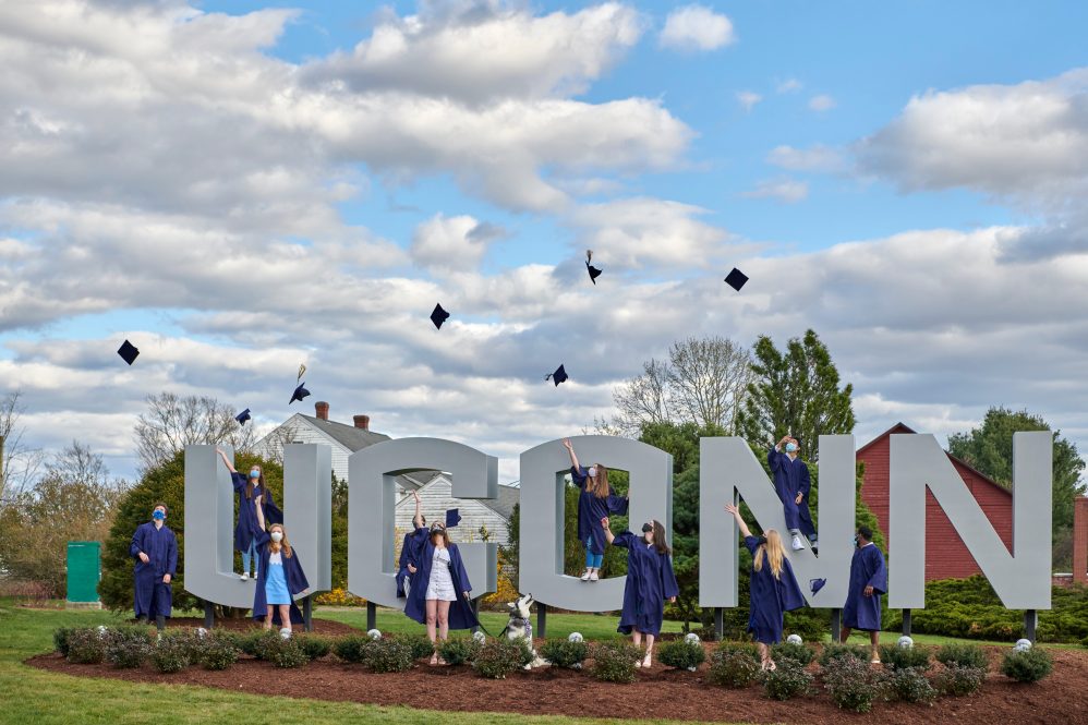 A group of graduating students toss their caps into the air while standing in front of the UConn gateway sign on April 28, 2020. The students are wearing facemasks due to the COVID-19 corona virus.