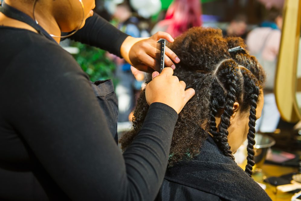 A close up of a Black hair stylist braiding a Black woman's hair. Connecticut recently outlawed workplace discrimination based on hairstyle, which UConn alumni and faculty members say is long overdue, especially for Black and Latino workers.