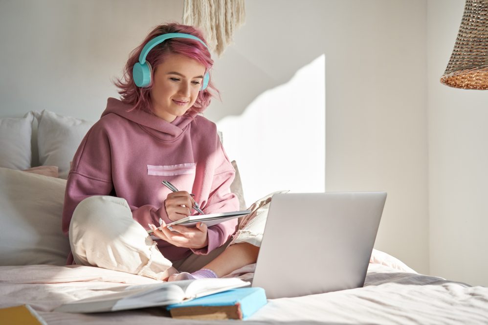 A young woman with headphones sits on her bed in front of a laptop during an online course. For students with disabilities, the shift to remote learning during the pandemic has brought challenges, but also benefits.