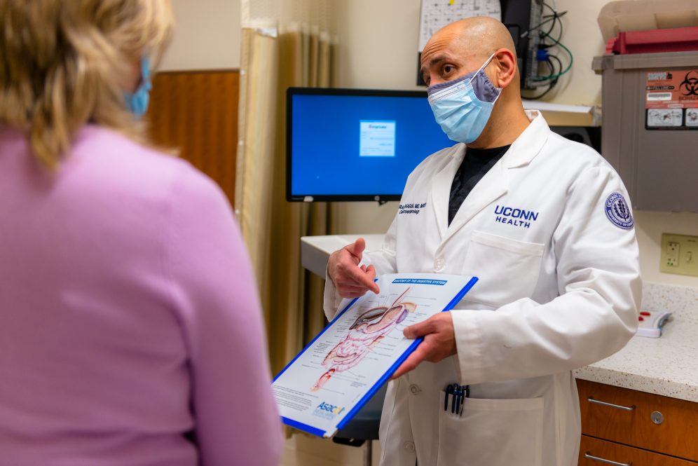 A doctor holding a clipboard with an illustration of the digestive system speaks to a patient in a medical office