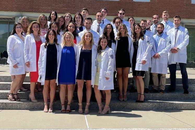 Several smiling students stand outside wearing white clinical coats